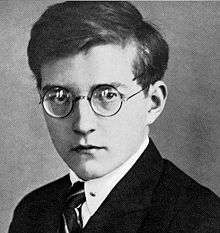 A young man wearing round-framed glasses, a dark jacket, tie and dress shirt