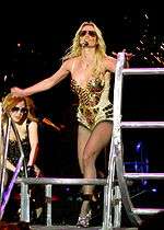 A female blond performer. She is wearing a sparkly black bodysuit and sunglasses. She also has a headset in her head. She is grabbing the bar of a jungle gym. In her right, a female is wearing sunglasses and a black corset with metal pieces.