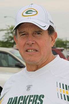 Head and shoulders photograph of Capers wearing a white Green Bay Packers t-shirt and white Packers baseball cap