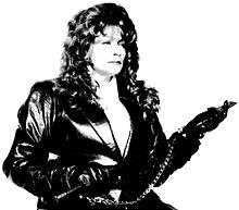 A black-and-white photograph of a woman with long, wavy hair wearing a black leather jacket, black leather gloves, and handcuffs