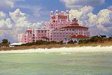 A pink, multi-floor hotel stands out above a calm sea and a windswept beach.