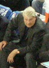 An older man, wearing a black suit, is sitting on the side of a basketball court.