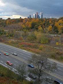A photo of a freeway in a ravine in the autumn, taken from atop the edge of the ravine. Projecting above the forest is the top of several skyscrapers and the CN Tower.