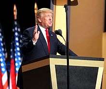 Trump standing behind a wooden, inverted-pyramid-shaped lectern with black paneling. He is speaking into a microphone, with an American flag hanging on a pole behind him. He is accepting the Republican nomination at the 2016 Republican National Convention in July 2016.