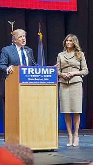 Donald and Melania Trump standing behind a blond-wood podium with the words "TRUMP", "TEXT 'TRUMP' TO 88022", "MANCHESTER, New Hampshire", and "MAKE AMERICA GREAT AGAIN" printed in white-on-blue text from top to bottom. Donald is to the left, behind the actual podium. Melania is about three feet to his left.