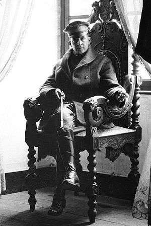 A man sits in an ornate chair. He is wearing a peaked cap, greatcoat and riding boots and holding a riding crop.