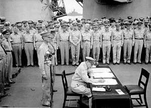 MacArthur is seated a small desk, writing. Two men in uniform stand behind him. A large crowd of men in uniform look on.