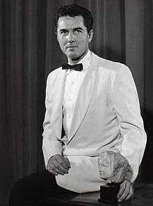 A man with dark hair, wearing a white suit including a black bow tie and pants, also holding an art piece.