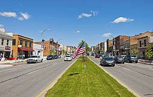 A wide street with two-lane roadways in either direction and older commercial buildings on either side, seen from a grassy median with the American flag flying from a low post in the center