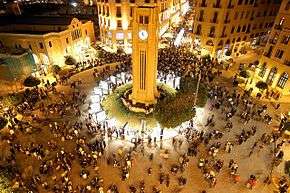 Bird's-eye view of people gathering around a clocktower at the center of a street light-lit square at night.