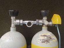 The top of a twin set of steel 7 litre cylinders shows a pair of Dräger vertical spindle taper thread valves with rubber knobs and DIN outlets linked by a single DIN centre-outlet manifold. The left side cylinder has a reserve lever with operating rod and a yellow plastic guard over the lever to reduce the risk of it being inadvertently opened by bumping against the surroundings