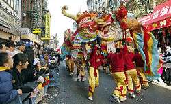 Several men in red and yellow outfits carry a colorful paper dragon in the street while onlookers watch behind police barriers.