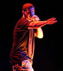 An illuminated man wearing a black T-shirt raps into a microphone.