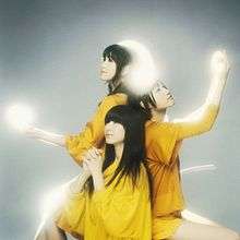 Three Japanese women (members from Perfume) sitting down, with yellow-ish lights circulating around them.