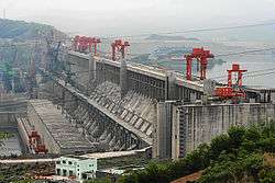 Aerial view of a huge concrete structure located between the mountains, with buildings, power lines and construction and lifting cranes