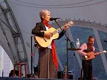 Baez plays outdoors in brown wide-leg pants, white top, brown waistcoat, blue pearls, and a long orange neck scarf. To her left, a male accompanist in a vest plays a small wooden cigar-box-style guitar