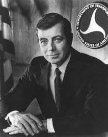 Black-and-white photo of a man wearing a suit sitting at a desk with his hands folded on it and the DOT logo and US flag behind him