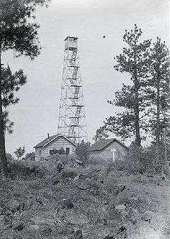 A four-legged tower with a small at the top, next to two one-story buildings. The tower is four stories tall. Trees are at either side, and in the foreground there are rocks, some vegetation, and a rough trail.