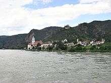 A view of Dürenstein from the river: a small town sits between steep mountains and a wide river. The church tower rises above the several dozen houses. On the mountain above, a ruined castle dominates the sky line.