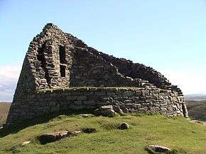 The ruins of a stone building set on a grassy knoll with blue skies above. The building is circular  in outline and all that remains of the structure are the double-skinned walls that rise to two stories in places.