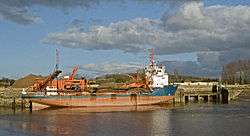 A cargo boat moored at a wharf with cranes and others machines. To the right is a metal gate opening to the water which flows past the boat.