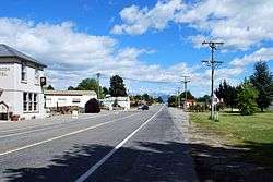 A straight road, divided by painted lines, passing through a town. On the left, the street is lined a pub and some widely separated shops. On the right, the road is lined by a grass verge with some small buildings in the middle distance. A black utility (car with a tray) is driving on the left away from the camera. A mountain range with a small covering of snow is in the background.