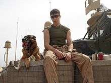 A black and tan German Shepherd dog sits next to a Marine atop a HECO barrier with a MTVR in the background. Lex is holding a red Kong toy in his mouth while Lee is wearing sunglasses and fire-resistant coveralls open to his waist.