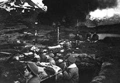 alt=Historic photograph of U.S. Marines in defensive trenches during the Japanese attacks of 1942, while fuel tanks burn in the background.