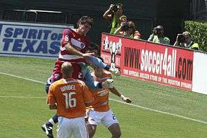 A Dallas FC player jumping in the air versus two Houston defenders and their goalkeeper