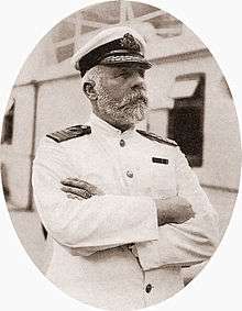 Photograph of a bearded man wearing a white captain's uniform, standing on a ship with his arms crossed