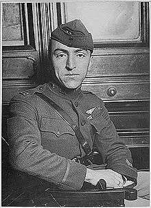 Head and torso of a man turned in his chair, his right arm lying stiffly on the arm rest in front of him. He is wearing a military uniform with a strap diagonally across the chest, a winged emblem on his left breast, and a garrison cap.