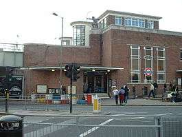 A plain red brick station entrance building. A statue of a knelling archer sits on a stone capped wall
