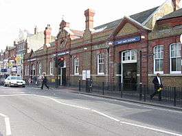 A red- and brown-bricked building with a blue sign reading "EAST HAM STATION" in white letters and people walking in front all under a white sky