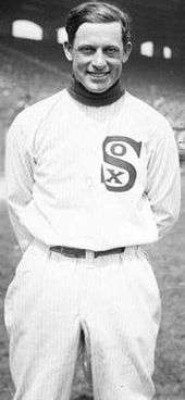 A man in a white baseball uniform smiles at the camera. He is shown from the knees up. His uniform shows the word "Sox", with a small "O" and a small "X" inside the vertical large letter "S". His hands are folded behind his back.