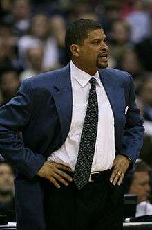 A man coaching from the sidelines of a basketball game. He is wearing a blue jacket and a white shirt with a dark, patterned tie.
