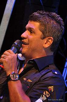 A man holding a microphone, wearing a black shirt and looking left.