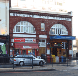 A red-bricked building with a white sign reading "EDGWARE ROAD STATION" in red letters with a car driving in the foreground