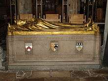 A stone rectangular monument, with the golden figure of a man dressed in robes and wearing a mitre lying on top, with his hands together in a gesture of prayer. The words "Edward Stuart Talbot" on the side in gold, with three shields bearing coats of arms below.  At the bottom, in gold: beneath the first shield, "1870 Keble 1888"; beneath the second, "1834–1944"; beneath the third, "1889 Leeds 1895"