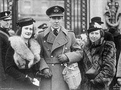 Photograph of a man with two women. The man is in the centre, wearing military uniform, a cap and a large coat, with a bag over his shoulder. He is holding the hand over the younger woman on his right. Both women are dressed formally, with hats and fur coats.