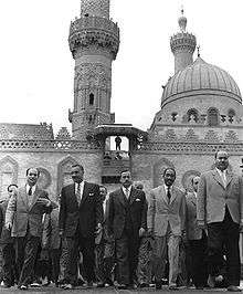 Several men walking forward, side-by-side. There are five men in the forefront, all wearing suits and ties. In the background is an ornate building with two minarets and a dome.