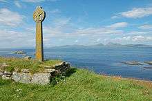 A stone cross in the Celtic style sits in a grassy field on an overgrown stone plinth. Small rocky reefs lie in the sea beyond, and there are high green hills in the distance.