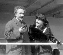 Einstein, looking relaxed and holding a pipe, stands next to a smiling, well-dressed Elsa who is wearing a fancy hat and fur wrap. She is looking at him.