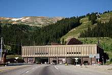 Automobiles are driving on a road leading to one of two openings in a building against a mountain. Letters above each opening read "Johnson Tunnel 1979" and "Eisenhower Tunnel 1973". On the roof of the building, large ventilation hoods are visible.
