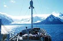  On-board view towards the bow of a small boat with a mast. Beyond the bow is a shoreline of snowy mountains.