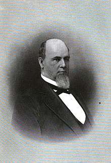 A man, balding on top with dark hair in the back and a gray beard, wearing a black jacket and white shirt