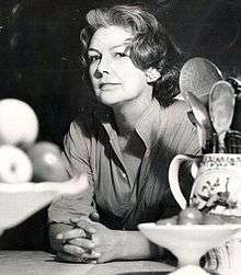 middle aged woman with dark, greying, hair; she is at a kitchen table, looking towards the camera