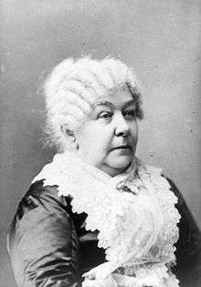 A monochrome photograph portrait of an elderly woman shown from the elbow up, turned somewhat to the right, a white lace apron covering a dark shiny fabric dress, the white hair in front-to-back rows of ringlets
