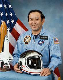 man in fight suit holding helmet, space shuttle model and American flag in background