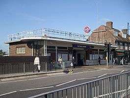 A brown-bricked building with a blue sign reading "ELM PARK STATION" in white letters and people walking in front all under a blue sky