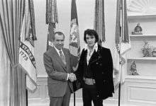 A mutton-chopped Presley, wearing a long velour jacket and a giant buckle like that of a boxing championship belt, shakes hands with a balding man wearing a suit and tie. They are facing camera and smiling. Five flags hang from poles directly behind them.